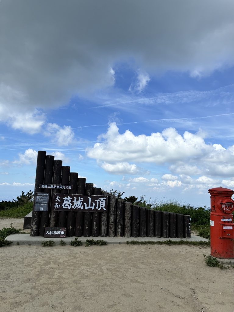 「大和葛城山頂」の標識と赤いポスト、背後に入道雲